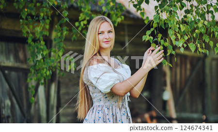 Young blonde female in traditional slavic attire caressing birch foliage within serene rural landscape Young blonde female in traditional slavic attire caressing birch foliage within serene rural landscape 124674341