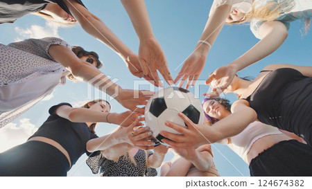 Group of young women forming a circle around a soccer ball with their hands, viewed from a low angle. 124674382