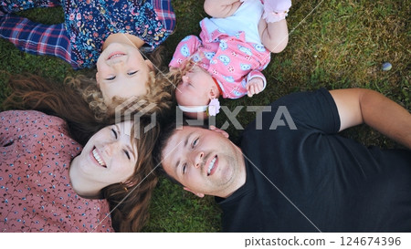 Smiling parents relaxing on verdant lawn, bonding with two young daughters and infant, embodying warmth and familial connection 124674396