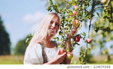Blonde woman picking fresh apples from sunlit branches, experiencing bountiful summer orchard harvest with joyful enthusiasm Blonde woman picking fresh apples from sunlit branches, experiencing bountiful summer orchard harvest with joyful enthusiasm 124674399