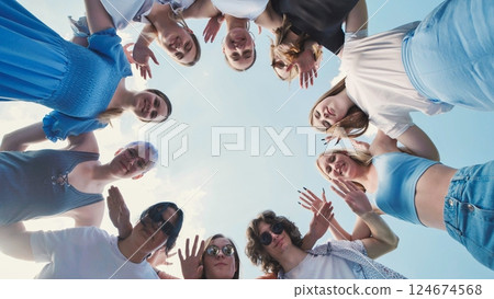 Young students gather in a circle, waving at the camera with a vibrant blue sky backdrop 124674568