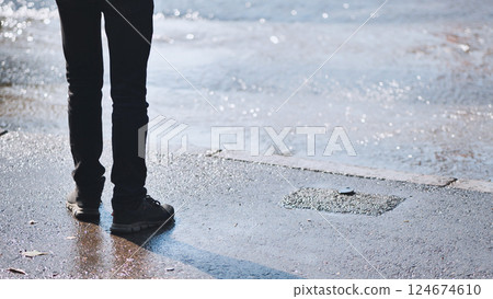 Close up of a man's legs and feet standing near a water leak on an asphalt road, observing the flowing water 124674610
