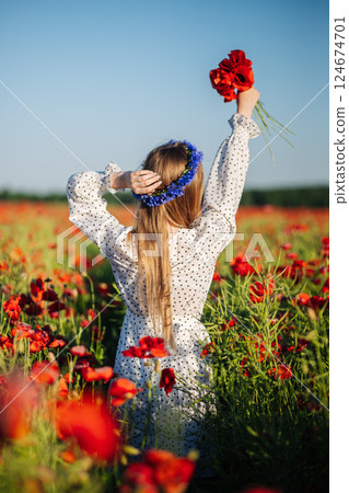 Blonde woman in white polka dot dress, flower crown, holding red poppies amid sunny wildflower meadow 124674701