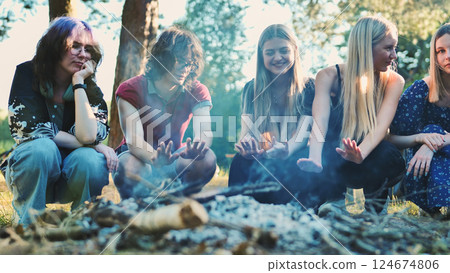 Friends warming hands around campfire, sharing camaraderie amid forest trees during evening outdoor gathering Friends warming hands around campfire, sharing camaraderie amid forest trees during evening outdoor gathering 124674806