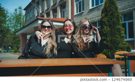 Three cheerful students in sunglasses sit at an outdoor school desk, smiling on a sunny day 124674860