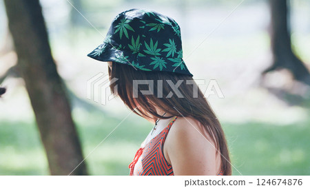 Young woman in cannabis leaf bucket hat relaxing during sunny park afternoon 124674876