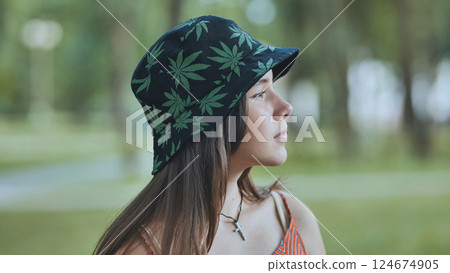 Young woman relaxing in park, sporting cannabis themed bucket hat on sunny day 124674905