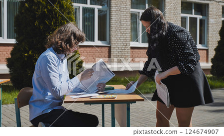 Two teachers are reviewing exam papers at a table outside a school building on a sunny day Two teachers are reviewing exam papers at a table outside a school building on a sunny day 124674907