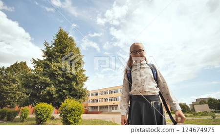 Cheerful schoolgirl with glasses and backpack standing near school, basking in sunlight under leafy trees and azure sky 124674975