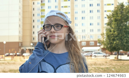 Young student with glasses, white baseball cap, texting on smartphone, smiling while walking urban street 124674983