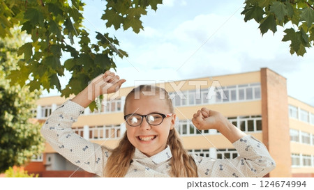 Happy school girl raising her arms in celebration in front of a school building, expressing joy and success Happy school girl raising her arms in celebration in front of a school building, expressing joy and success 124674994