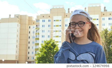 Cheerful schoolgirl with glasses and cap chatting on smartphone, standing near school building during bright summer afternoon Cheerful schoolgirl with glasses and cap chatting on smartphone, standing near school building during bright summer afternoon 124674998