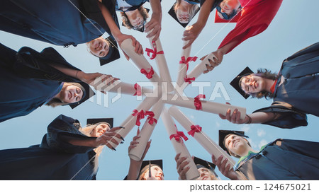 Group of cheerful graduates forming a circle, holding their diplomas together with pink ribbons, seen from a low angle 124675021