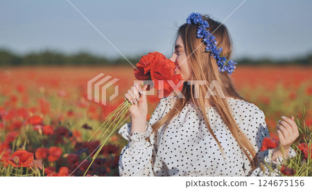 Blonde woman smiling, wearing blue flowers in hair, standing amid vibrant red poppy field and inhaling deeply near blooming blossoms Blonde woman smiling, wearing blue flowers in hair, standing amid vibrant red poppy field and inhaling deeply near blooming blossoms 124675156