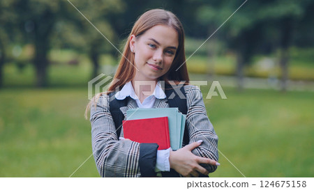 Portrait of a smiling schoolgirl holding books in a park, enjoying the back to school season 124675158