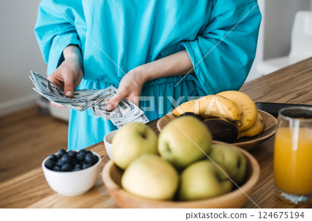 Woman counting US dollar bills next to fresh fruit and juice on a kitchen counter. Money management, personal finance, budgeting for wellness, healthy lifestyle economics 124675194