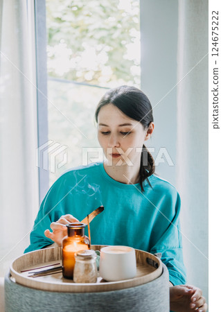 Young woman burning palo santo near a tray with candles and tea in a calm home setting. Sensory mindfulness for daily balance, palo santo ritual, inner calm, grounding therapy 124675222