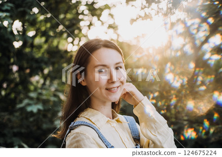 Young woman with fair skin and brown hair smiling gently in a sunlit outdoor setting. Positive mindset, emotional wellness, self-acceptance, personal growth 124675224