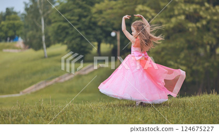 Young woman dancing joyfully, wearing pink dress, twirling gracefully in lush green park during bright summer afternoon 124675227