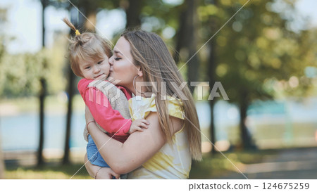 Loving mom tenderly kissing baby girl while cuddling outdoors, green park background 124675259