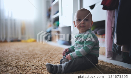 Toddler wearing blue pajamas, sitting on soft bedroom carpet, playing contentedly near wooden closet during cold winter morning 124675295