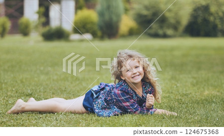 Happy young girl with curly blonde hair relaxing on a green lawn, enjoying a sunny family day in the backyard Happy young girl with curly blonde hair relaxing on a green lawn, enjoying a sunny family day in the backyard 124675368