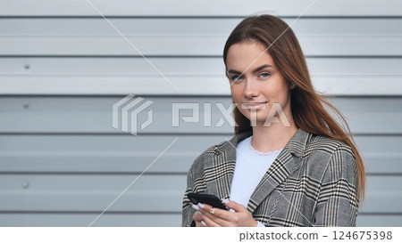 Professional businesswoman checking smartphone, leaning against sleek grey wall, embodying contemporary urban workplace style 124675398