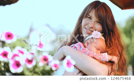Smiling mother holding her sleeping newborn baby girl in a garden with pink and white flowers 124675468