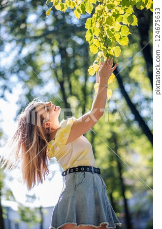 Teenage girl with flowing blond hair gently touching green tree leaves during sunny park afternoon Teenage girl with flowing blond hair gently touching green tree leaves during sunny park afternoon 124675653