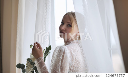 Elegant blonde bride wearing white gown, holding floral bouquet, gazing softly over shoulder near bright window Elegant blonde bride wearing white gown, holding floral bouquet, gazing softly over shoulder near bright window 124675757