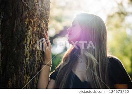 Blonde teenage student with long hair wearing black dress leaning against tree trunk enjoying nature in summer 124675760