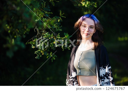Stylish woman with purple hair and glasses leaning against tree, basking in warm summer sunlight amid green park backdrop 124675767