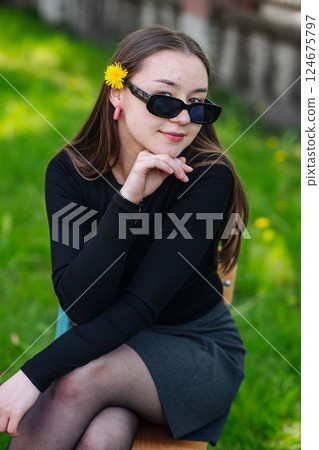 Stylish student with sunglasses and a dandelion in her hair relaxing on a bench in a lush park on a school day Stylish student with sunglasses and a dandelion in her hair relaxing on a bench in a lush park on a school day 124675797