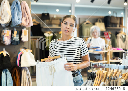 Young female buyer in store has removed hanger with trousers from window and is considering product. 124677648