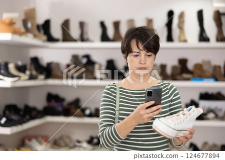 Female customer examines shoes in store and scans label with phone camera. Female customer examines shoes in store and scans label with phone camera. 124677894