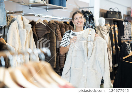 Girl stopped near showcase with outerwear, holds fur coat hanger in hands and examines product 124677945