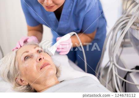Young female doctor, dermatologist using ultrasound cavitation machine for improvement of skin condition of old female in clinic 124678112