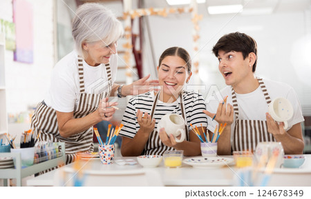 Cheerful older woman with young adult children painting pottery in family workshop 124678349