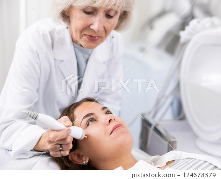 Young woman receiving non-surgical rejuvenation procedure from senior cosmetologist, RF face lifting with use of electric device equipment in spa center Young woman receiving non-surgical rejuvenation procedure from senior cosmetologist, RF face lifting with use of electric device equipment in spa center 124678379