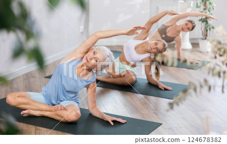 Three generation of women perform half-lotus pose of Ardha Padmasana with body tilted to side in gym 124678382