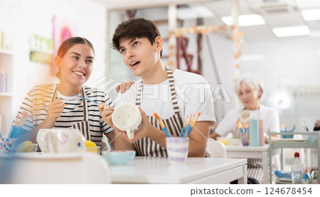 Cheerful young couple decorating ceramic pottery at table in workshop Cheerful young couple decorating ceramic pottery at table in workshop 124678454