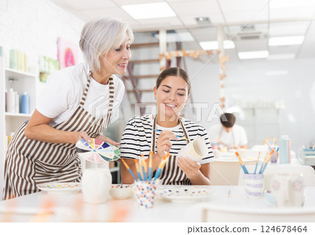 Elderly mother guiding adult daughter painting mugs in family pottery workshop Elderly mother guiding adult daughter painting mugs in family pottery workshop 124678464