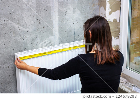 A woman is carefully measuring a radiator using a tape measure 124679298