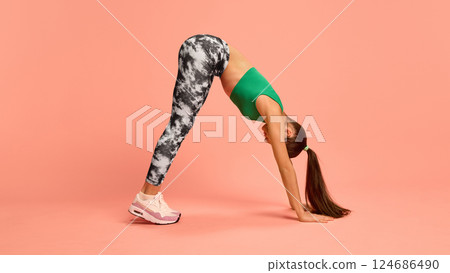 Fit girl in dynamic pose doing forward bend stretch, showing spine flexibility and full-body activation in black-white leggings and green crop top. 124686490
