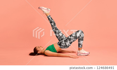 Athletic girl lying on floor with one leg raised, performing bridge exercise in stylish gym wear, building core strength and balance on pastel background. 124686491