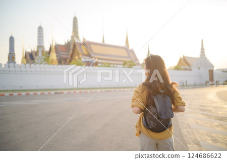 Traveler asian woman in her 30s, backpack slung over her shoulder, explores the intricate details of Wat Pra Kaew with childlike wonder. Sunlight dances on the golden rooftops. Traveler asian woman in her 30s, backpack slung over her shoulder, explores the intricate details of Wat Pra Kaew with childlike wonder. Sunlight dances on the golden rooftops. 124686622