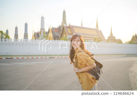 Traveler asian woman in her 30s, backpack slung over her shoulder, explores the intricate details of Wat Pra Kaew with childlike wonder. Sunlight dances on the golden rooftops. Traveler asian woman in her 30s, backpack slung over her shoulder, explores the intricate details of Wat Pra Kaew with childlike wonder. Sunlight dances on the golden rooftops. 124686624
