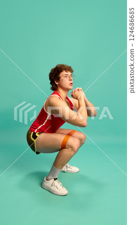 Focused man in red top squatting with resistance band on legs, engaging core and leg muscles against mint studio background. 124686685