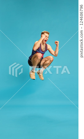 Sporty man in jump with strong expression, mid-air pose shows energy, power and confidence against blue background. 124686796