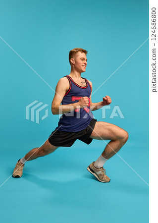 Sporty man doing side lunge with smile, dressed in shorts and tank top, showing strength and flexibility, against blue background. Sporty man doing side lunge with smile, dressed in shorts and tank top, showing strength and flexibility, against blue background. 124686800